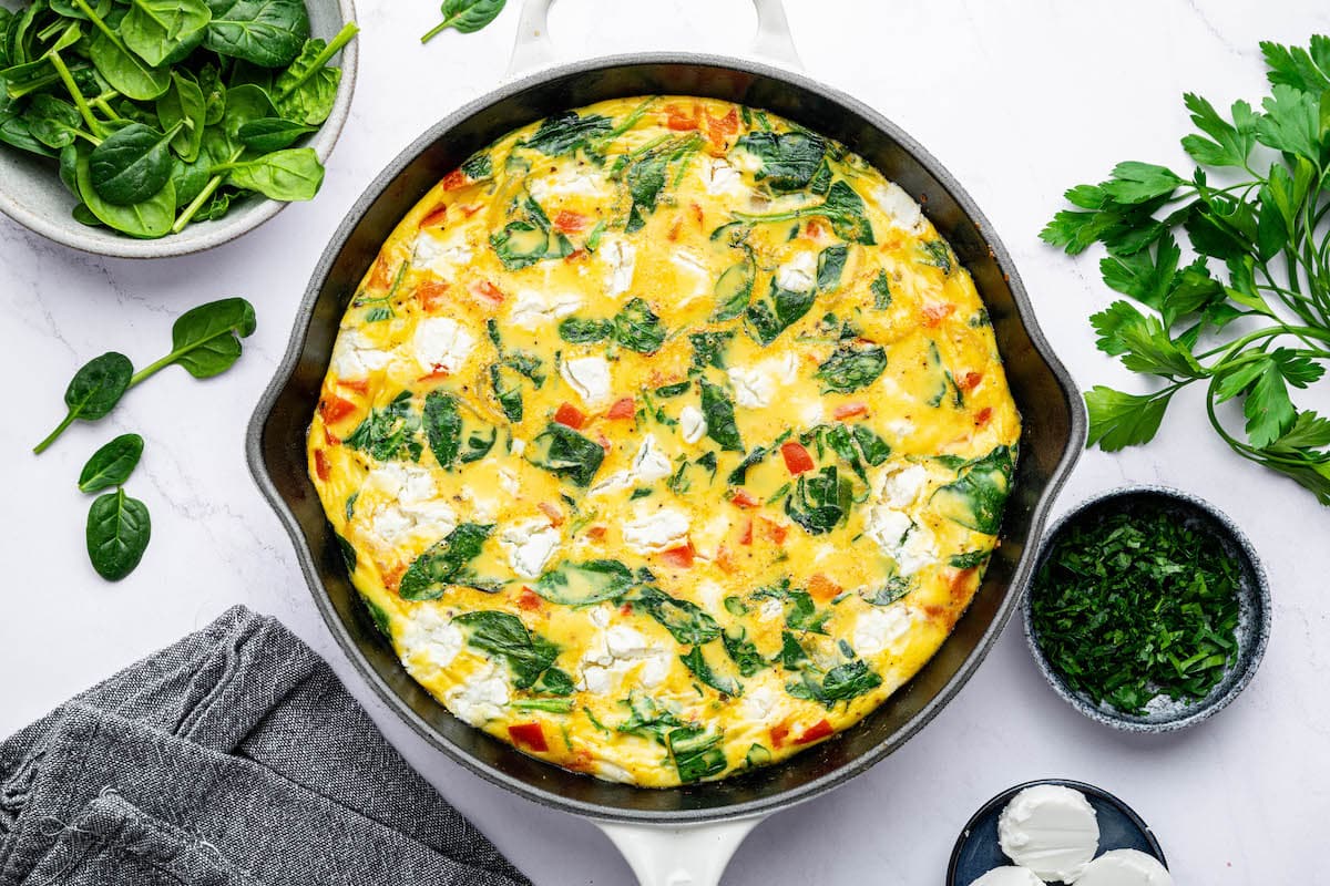 A cooked frittata in a white cast iron skillet. The frittata has red bell pepper, spinach, onion and goat cheese.