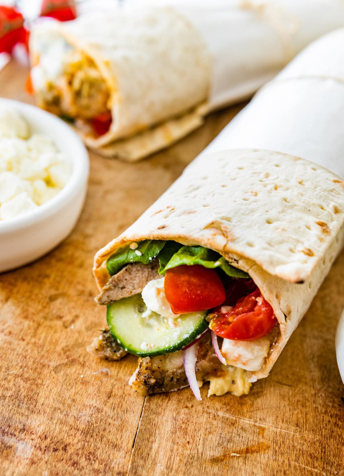 A Greek chicken wrap on a wooden cutting board. You can see the tomatoes, cucumber, red onion, chicken and lettuce inside the wrap.