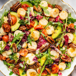 A large bowl of hearts of palm salad that has cherry tomatoes, arugula, avocado, pecans, and a honey balsamic dressing.