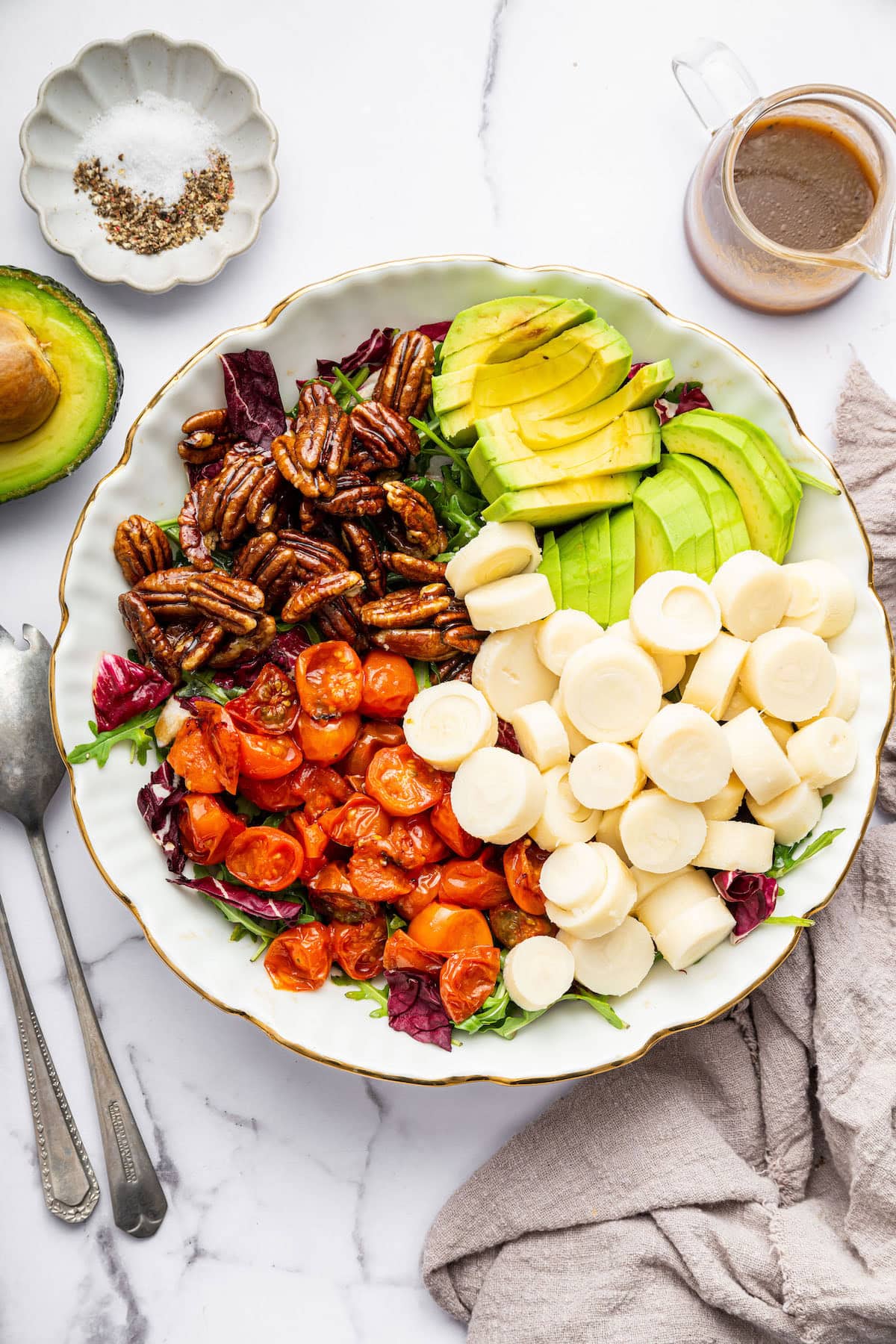 A large serving bowl filled with hearts of palm, roasted cherry tomatoes, avocado slices, and candied pecans arranged over a bed of mixed greens, with a jar of vinaigrette and small dish of salt and pepper nearby.
