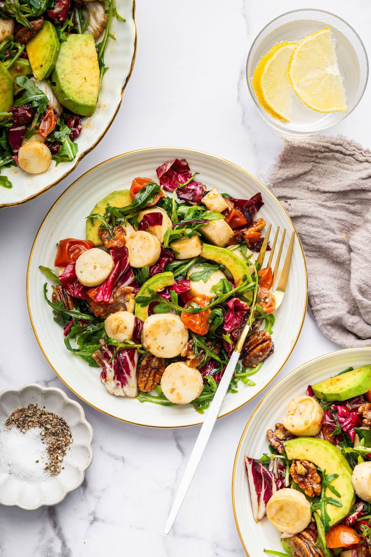 A plate of hearts of palm salad with avocado slices, roasted cherry tomatoes, radicchio, arugula, and candied pecans, served with a fork on a white plate.