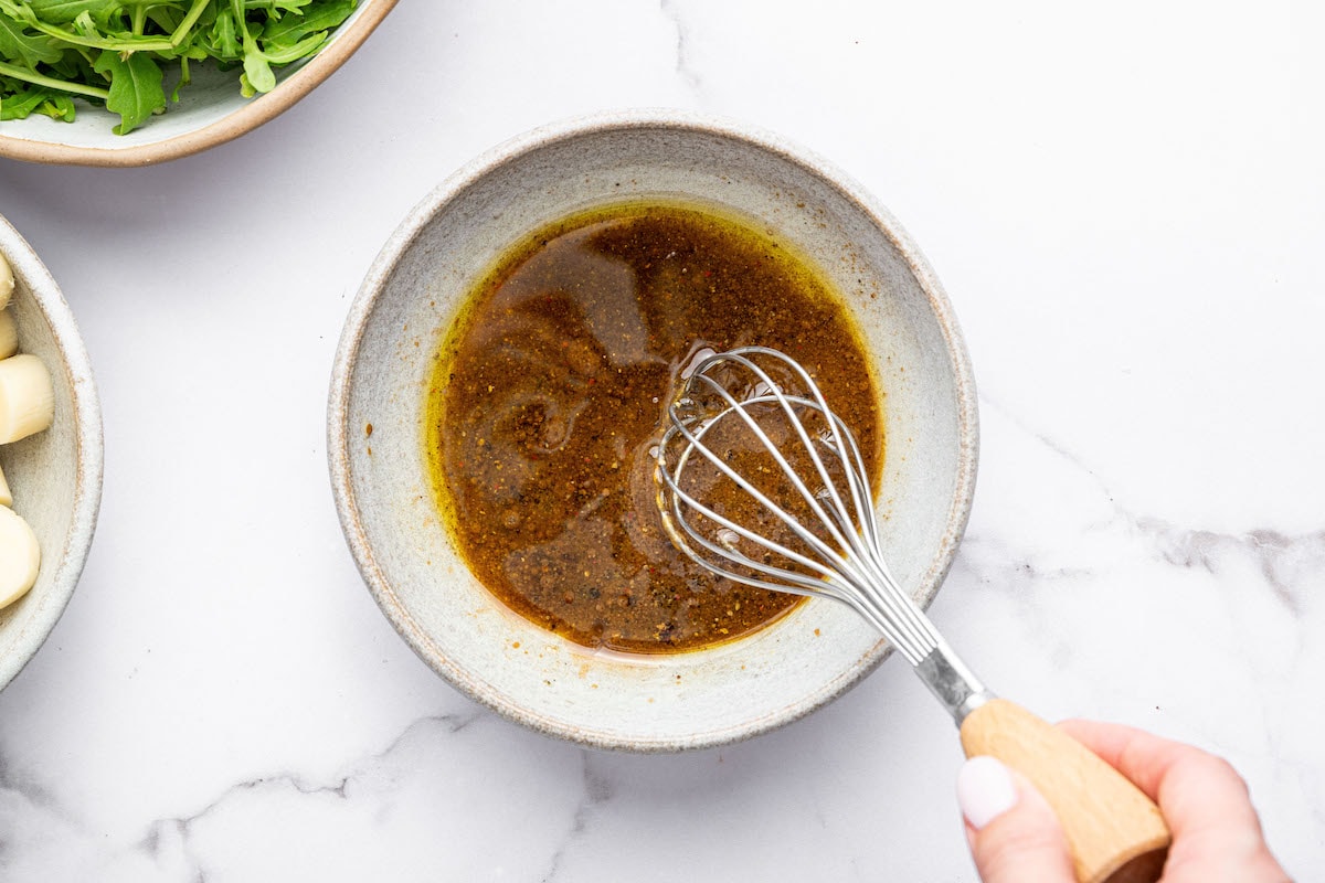 A small bowl of golden-brown vinaigrette dressing being whisked with a metal whisk by hand, surrounded by ingredients including fresh arugula and hearts of palm on a white marble surface.