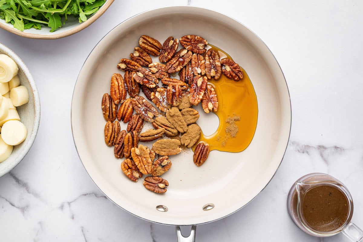 Pecans in a white skillet with maple syrup, cinnamon, and cayenne pepper, ready to be candied.