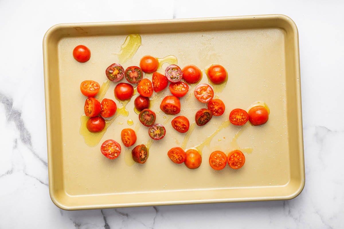 Halved cherry tomatoes drizzled with olive oil on a gold baking sheet, prepped for roasting.