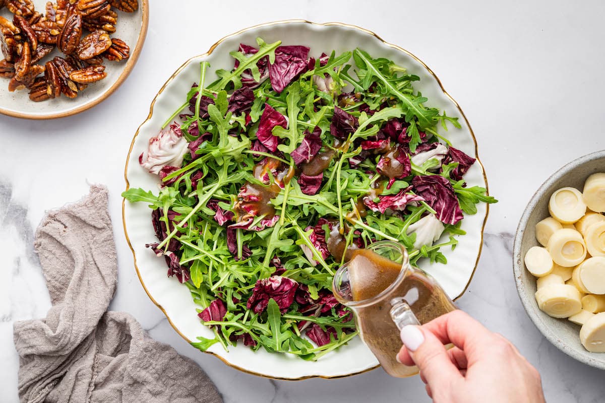 Hand pouring balsamic vinaigrette over a bowl of mixed greens with radicchio and arugula.