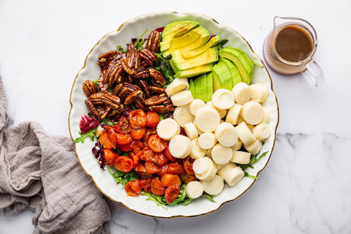 A large bowl of ingredients used in a hearts of palm salad before being mixed. This includes hearts of palm, cherry tomatoes, pecans, and avocado all on a bed of arugula.