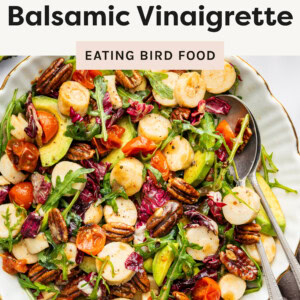 A vibrant hearts of palm salad served in a white dish with gold trim. The salad includes sliced hearts of palm, avocado slices, roasted cherry tomatoes, arugula, radicchio, and candied pecans, all tossed in a honey balsamic vinaigrette. A spoon and fork rest on the dish, with a halved avocado and a glass of water with lemon wedges nearby on a marble countertop.