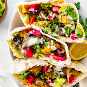 Three halves of chicken shawarma pita pockets stacked in a bowl, filled with veggies, olives, pickled turnips, and topped with yogurt sauce and fresh parsley.