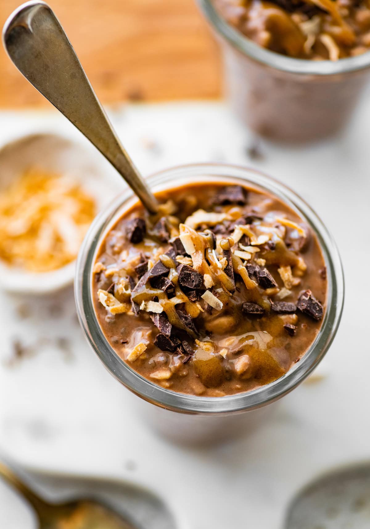 One glass jar filled with samoas overnight oats. There's a spoon in the oats. It's topped with chocolate chunks, date caramel and toasted coconut.