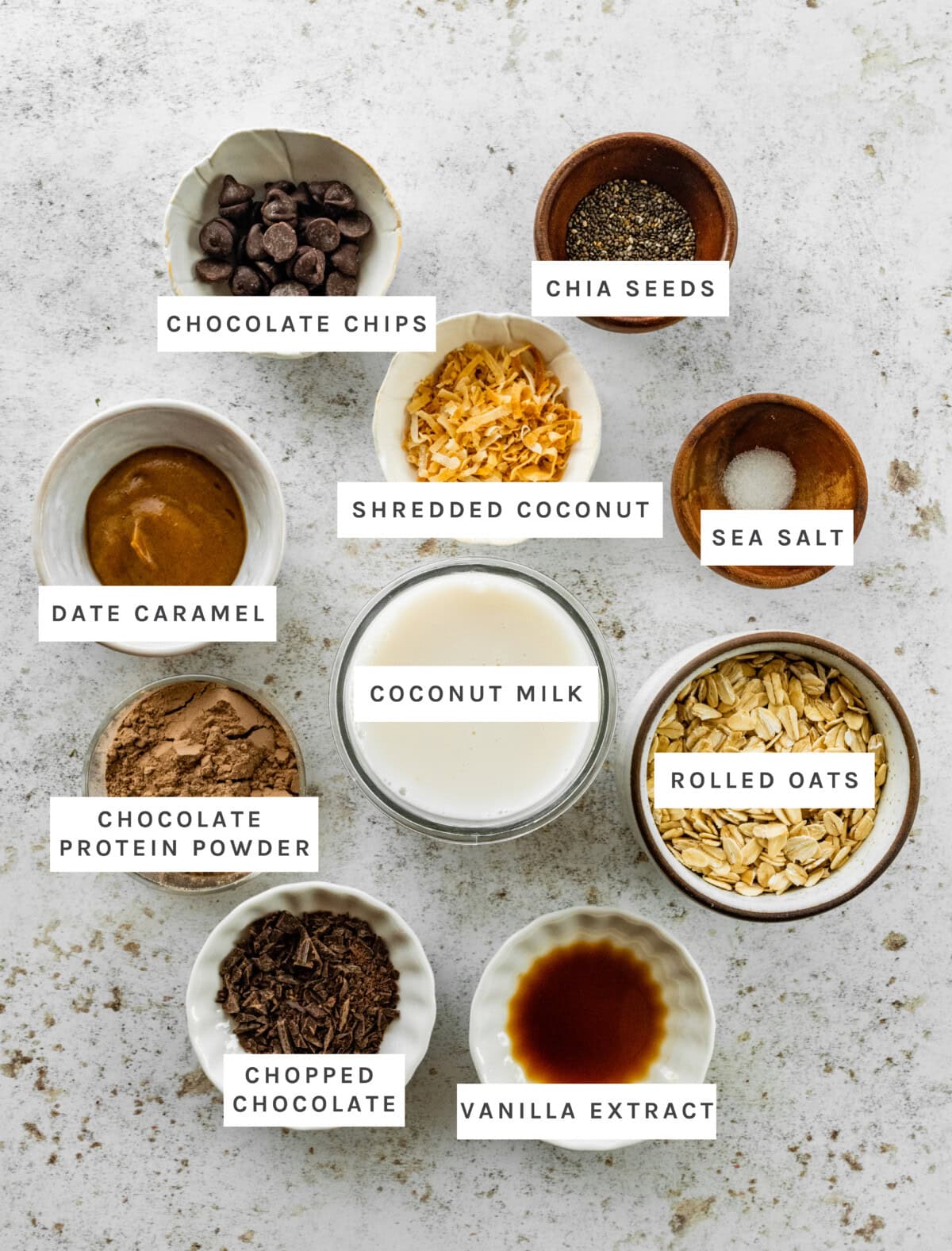 Ingredients measured out to make Samoas Overnight Oats: Chocolate chips, chia seeds, shredded coconut, sea salt, date caramel, coconut milk, rolled oats, chocolate protein powder, chopped chocolate, vanilla extract.
