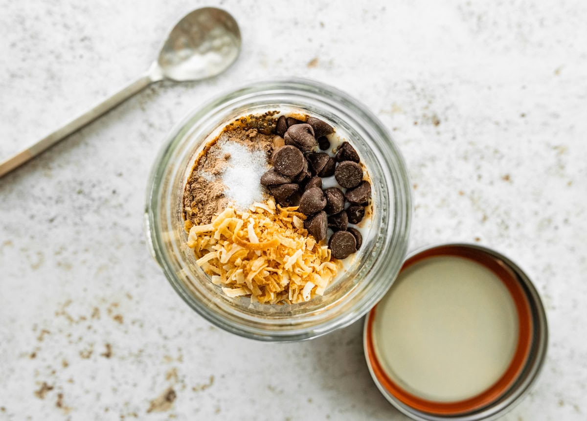 All the ingredients for the samoas overnight oats in a glass jar: protein powder, toasted coconut, salt, chocolate chips, oats, date caramel, milk and chia seeds.