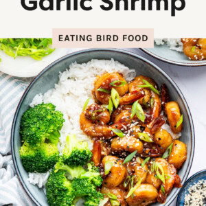 A bowl of honey garlic shrimp served over white rice, accompanied by steamed broccoli. The shrimp are coated in a glossy, caramelized sauce and garnished with sesame seeds and thinly sliced green onions. A second bowl with the same dish is partially visible in the background. A small dish of sesame seeds and a plate of additional green onions are placed nearby on a white surface with a striped cloth napkin.