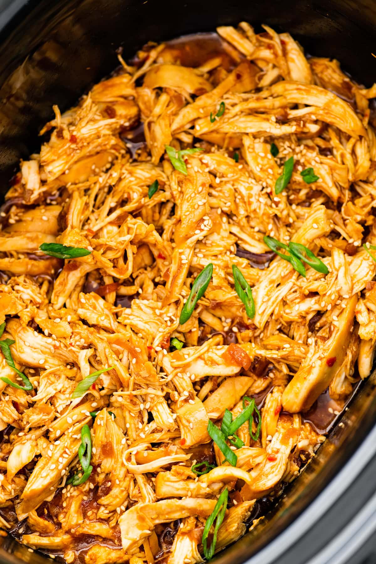 Shredded teriyaki chicken in a slow cooker. The chicken is topped with sesame seeds and green onions.