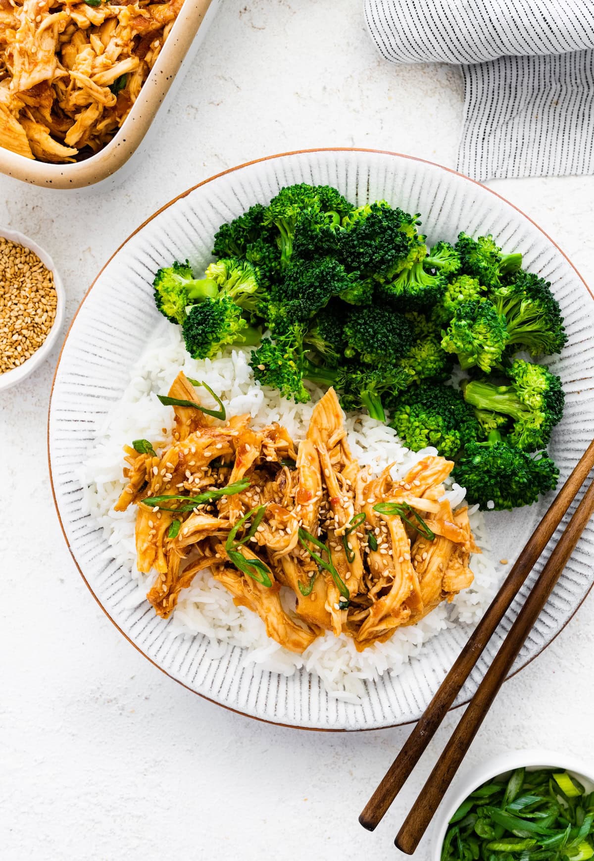 Slow cooker teriyaki chicken served on a plate with white rice and broccoli. There are chopsticks on the plate. The chicken is topped with sesame seeds and green onions.
