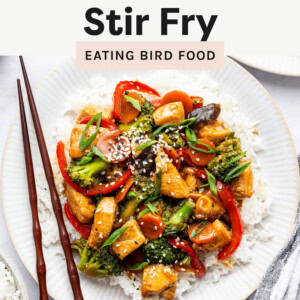 A plate of chicken stir fry served over white rice, garnished with sesame seeds and sliced green onions. The dish contains vibrant vegetables, including red bell peppers, broccoli, carrots, mushrooms, and onions, coated in a glossy, flavorful sauce. A pair of wooden chopsticks rests on the side of the plate, and a striped cloth napkin is partially visible in the corner. The background includes another plate of the dish and a bowl of rice.