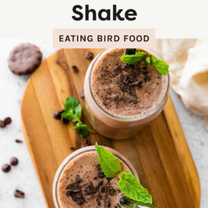 Thin Mint Protein Shake: An overhead view of two chocolate mint protein shakes in glass jars, topped with crushed chocolate cookies and fresh mint leaves. The shakes have a smooth, frothy texture and are served with green reusable straws. They are placed on a wooden board, surrounded by chocolate chips, cookie crumbs, and whole thin mint cookies, with a soft beige cloth in the background.