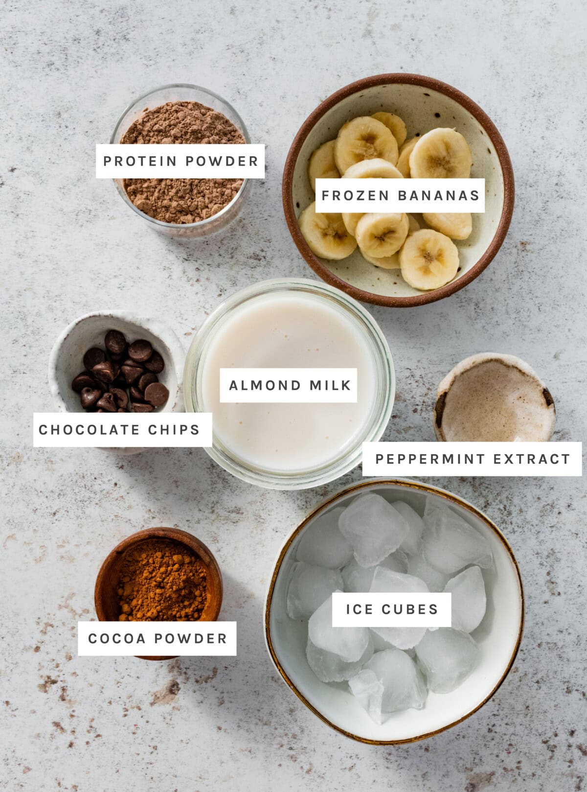 Ingredients measured out to make Thin Mint Protein Shake: Protein powder, frozen bananas, almond milk, peppermint extract, chocolate chips, cocoa powder, ice cubes.