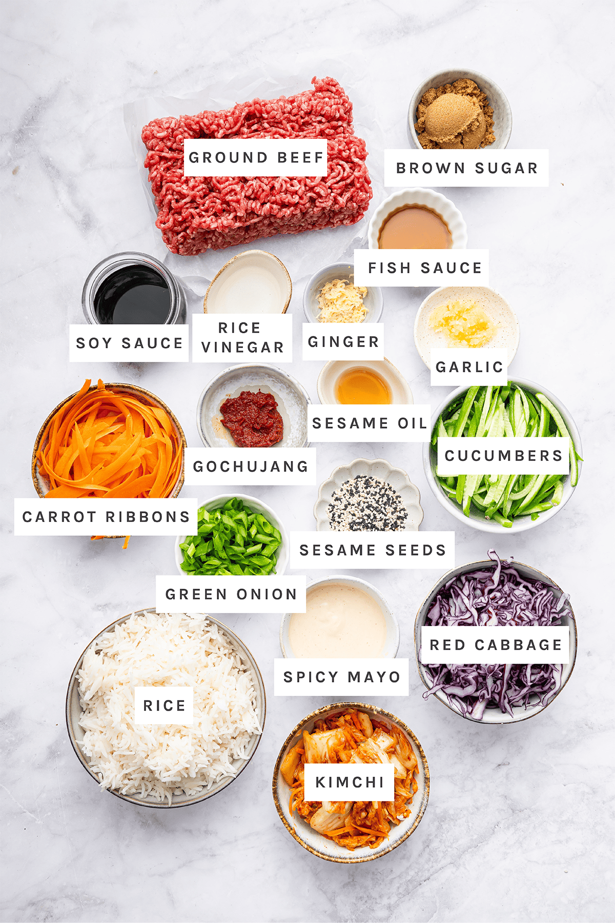 Ingredients measured out to make Korean Beef Bowl: Ground beef, brown sugar, fish sauce, soy sauce, rice vinegar, ginger, garlic, sesame oil, cucumbers, carrot ribbons, gochujang, sesame seeds, green onion, red cabbage, rice, spicy mayo, kimchi.