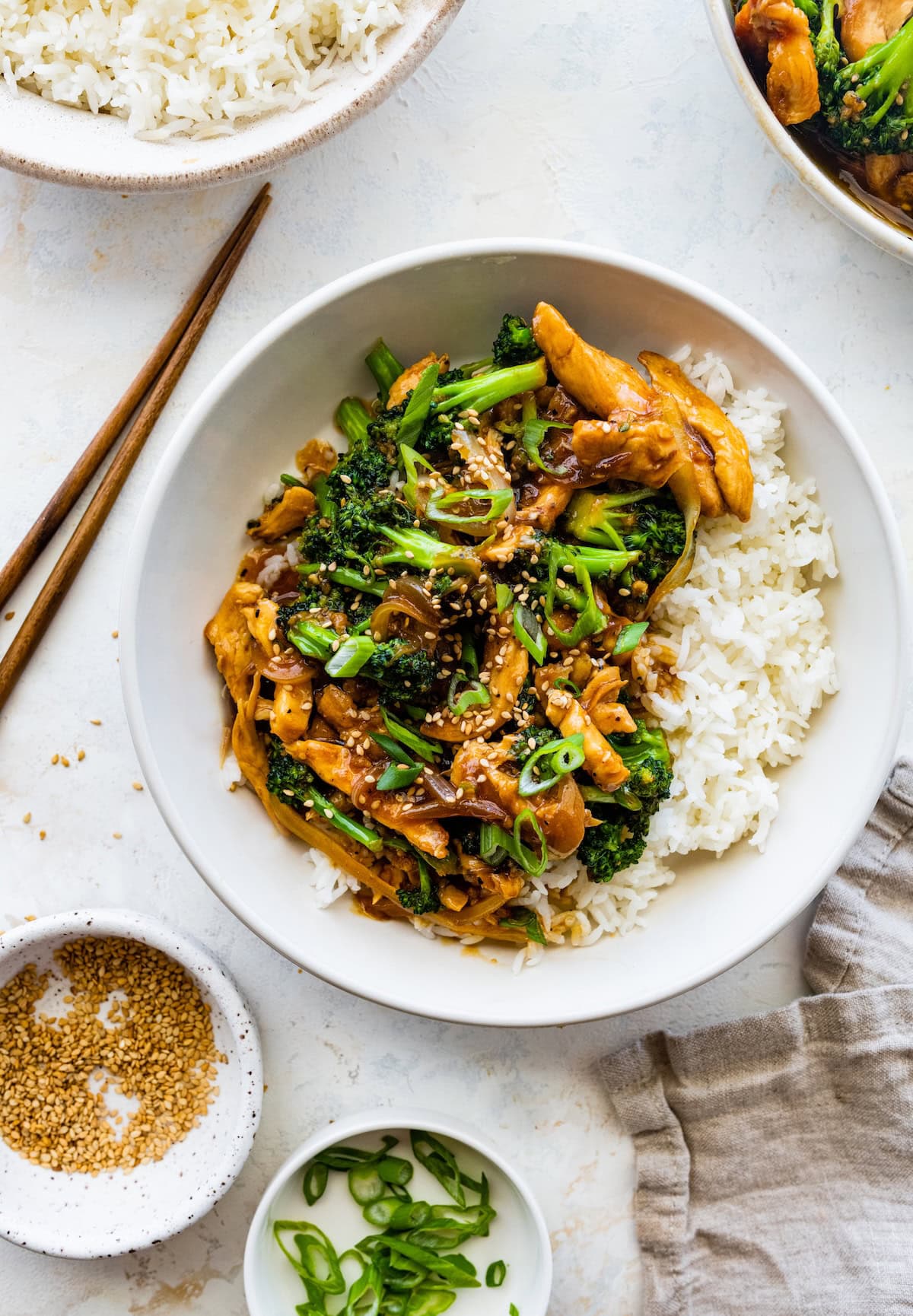 Chicken and broccoli stir fry in a white bowl, served with white rice and garnished with sesame seeds and green onions. Wooden chopsticks are next to the bowl.