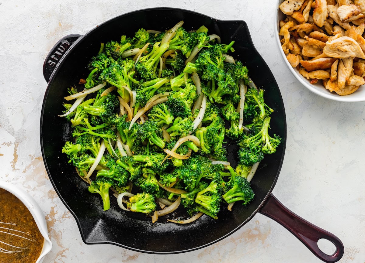 Cooked broccoli and yellow onions in a cast iron skillet next to a bowl of cooked chicken.