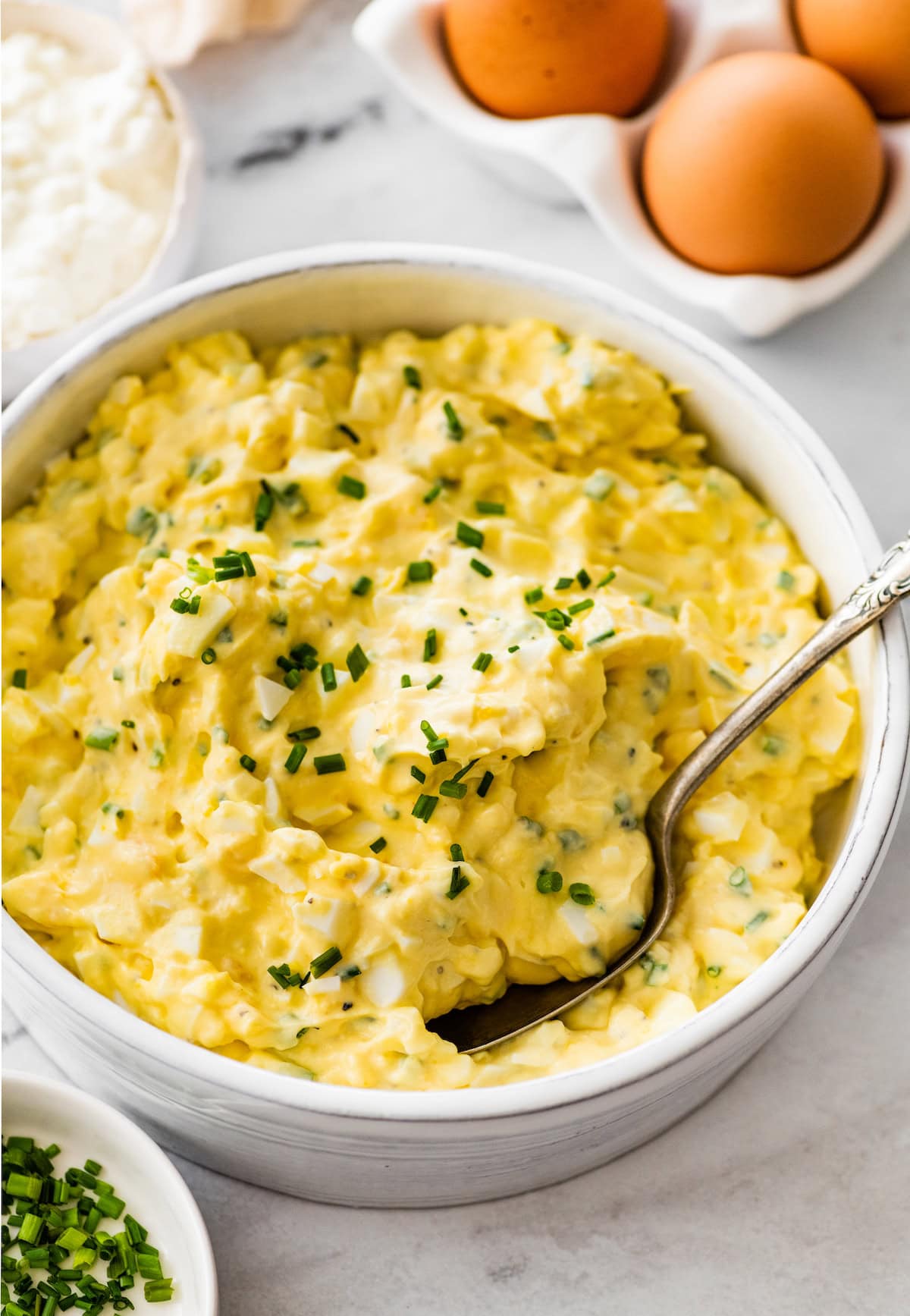 Cottage cheese egg salad served in a white ceramic bowl served with a silver spoon. Fresh chives are topping the egg salad.