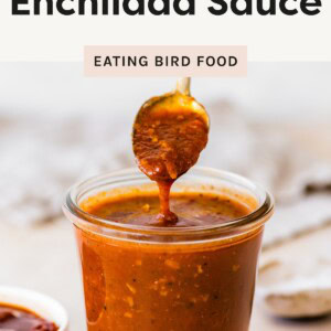 A spoonful of enchilada sauce drips back into a glass jar filled with the rich, red sauce; a small bowl of chipotle peppers sits nearby.