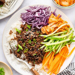 Korean beef served over white rice with fresh carrots, purple cabbage, cucumber and kimchi. The beef is topped with sesame seeds and green onions.