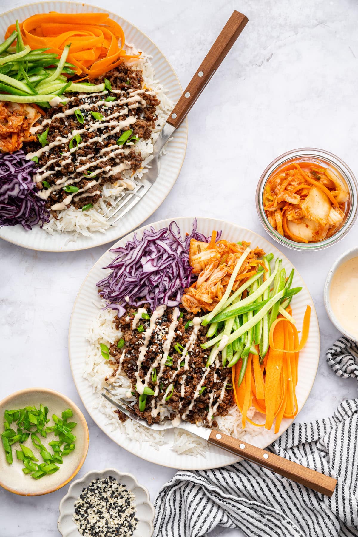 Two Korean beef bowls with white rice, fresh carrots, purple cabbage, cucumber and kimchi. The beef is topped with sesame seeds, green onions and a drizzle of spicy mayo.