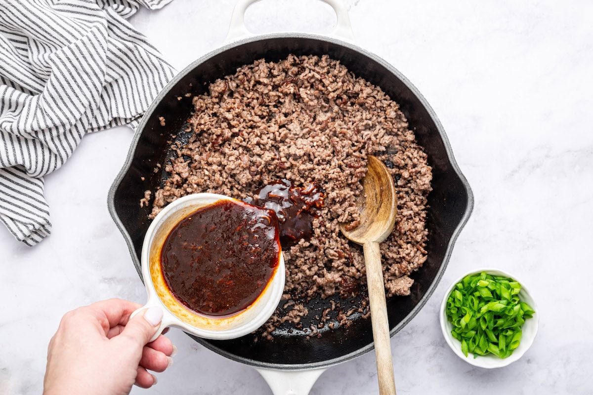 A woman's hand pouring the sauce mixture over the ground beef in a skillet. There is a wooden spoon in the skillet.