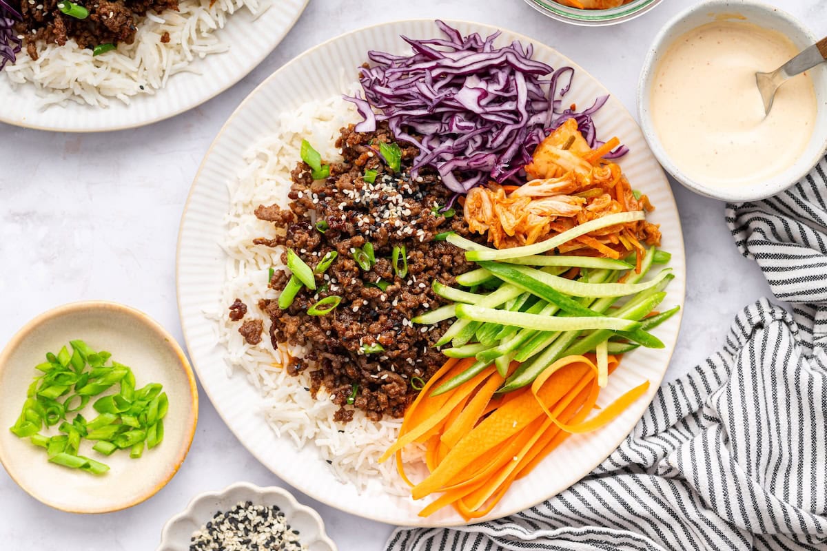Korean beef served over white rice with fresh carrots, purple cabbage, cucumber and kimchi. The beef is topped with sesame seeds and green onions.