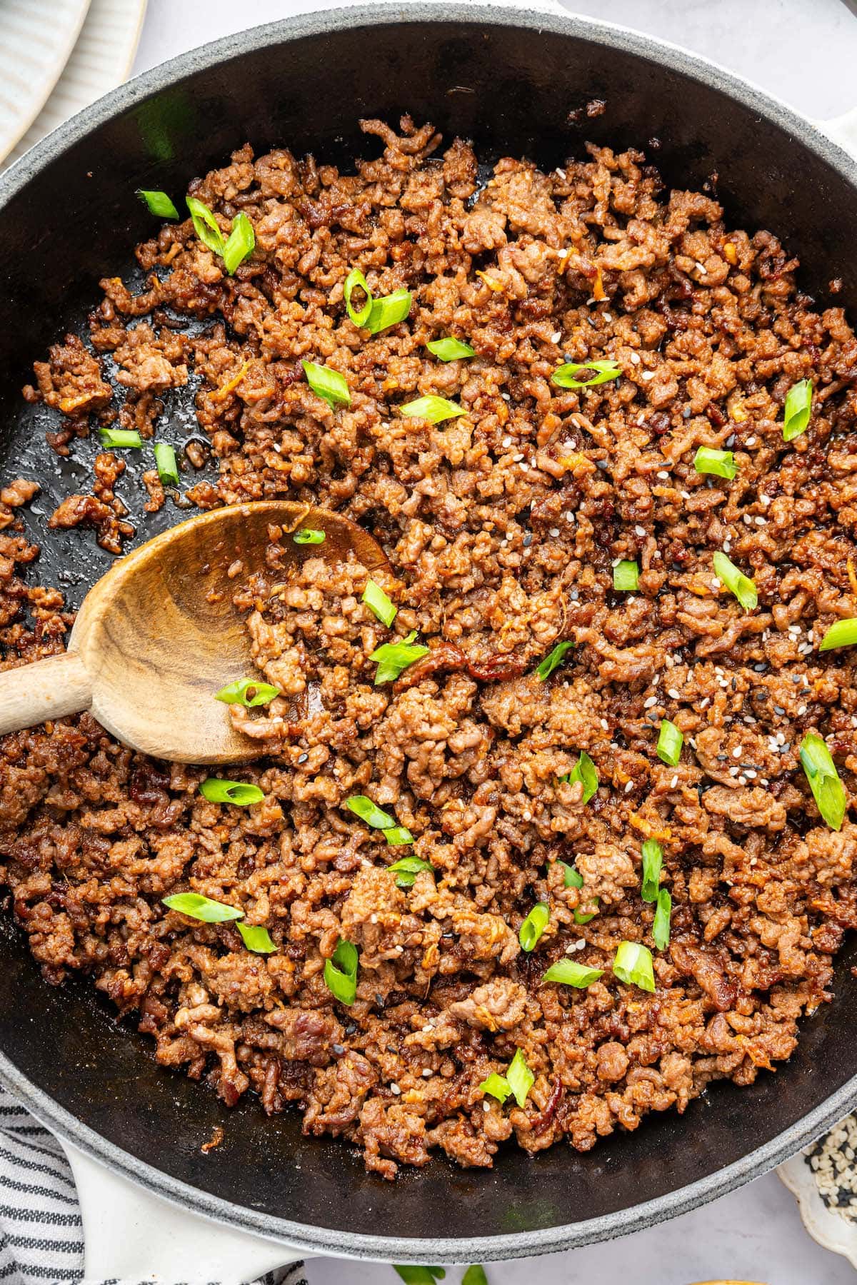 Korean beef mixture cooked in a skillet topped with green onion and sesame seeds. There is a wooden spoon in the skillet.