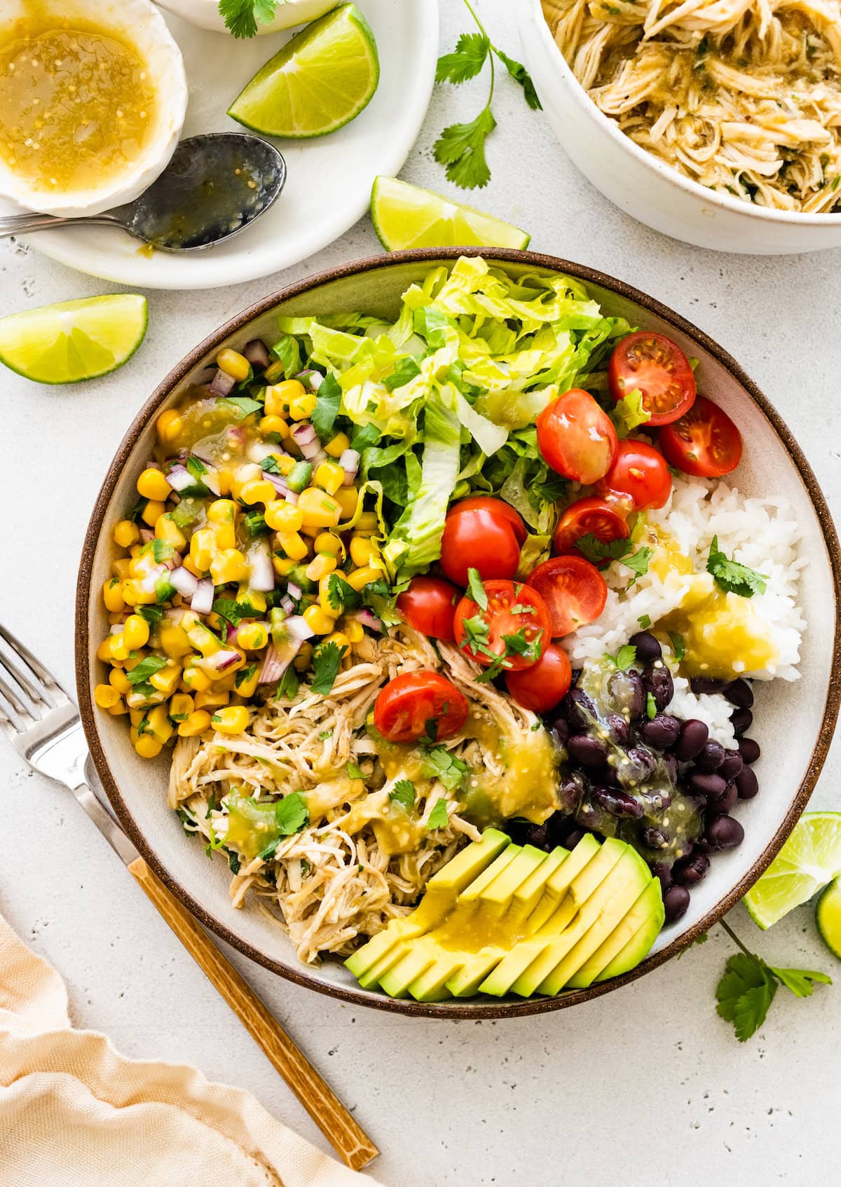 A bowl filled with shredded salsa verde chicken, avocado, black beans, white rice, cherry tomatoes, lettuce and a corn salad.