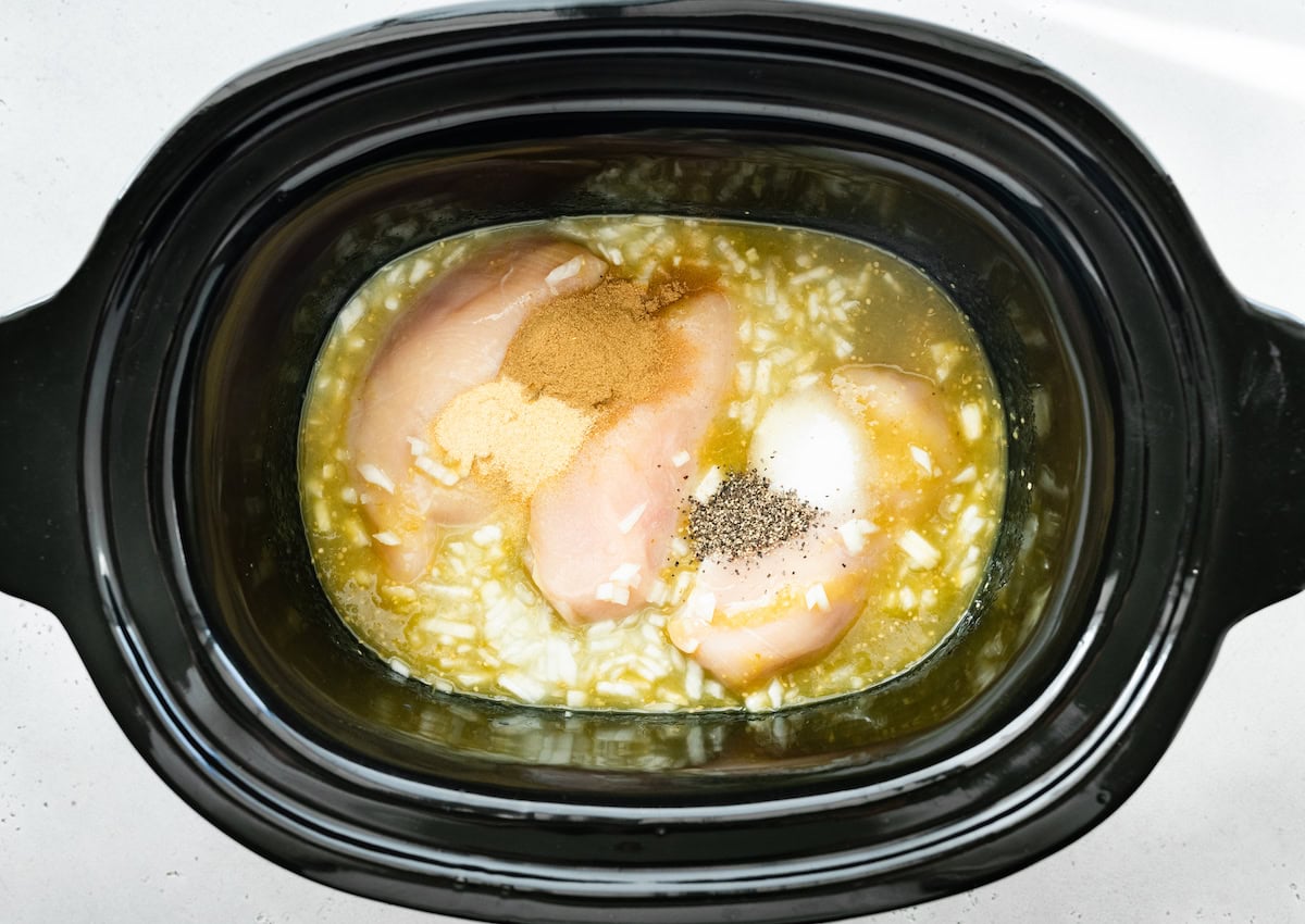 Three raw chicken breasts in a slow cooker with salsa verde, onion and spices.