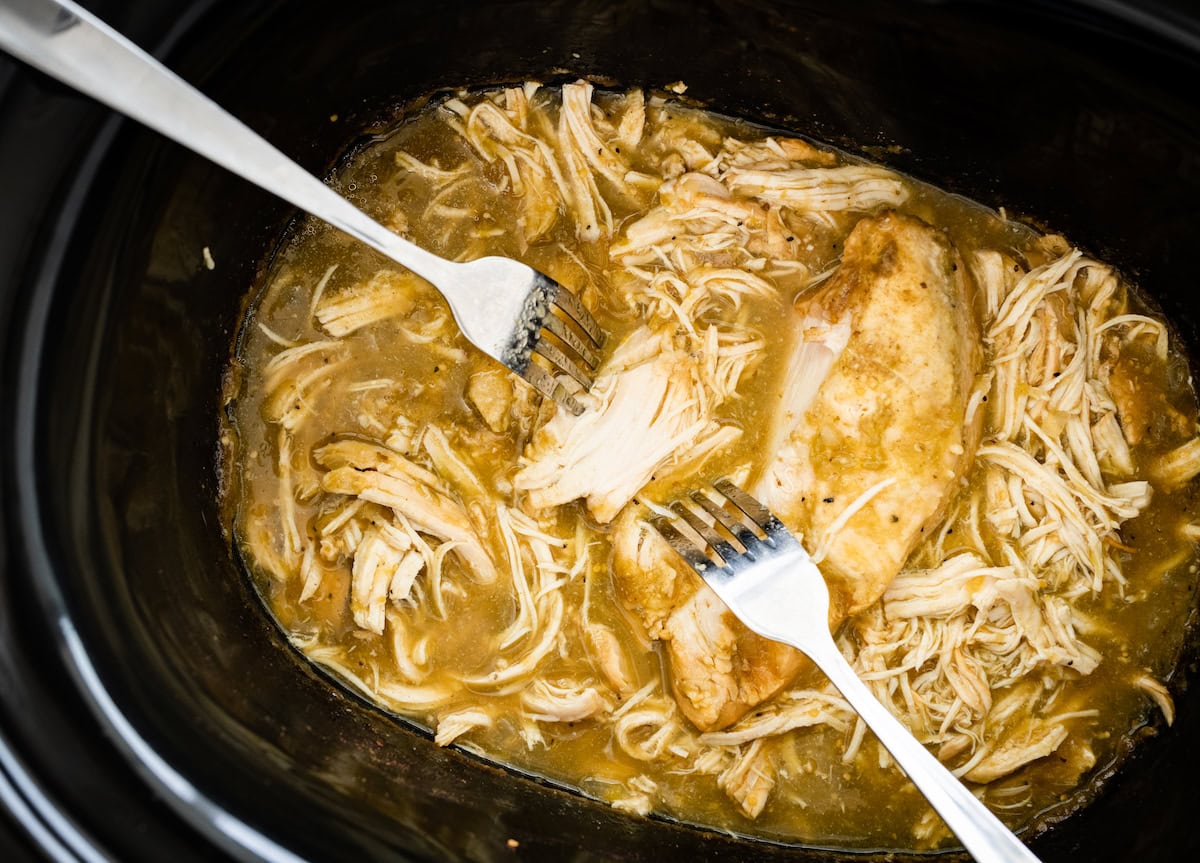 Two forks shredding the chicken breasts in the slow cooker with the salsa verde sauce.