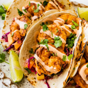 Three shrimp tacos in flour tortillas folded up on a serving platter. The tacos are topped with a mango cabbage slaw and a drizzle of spicy crema.