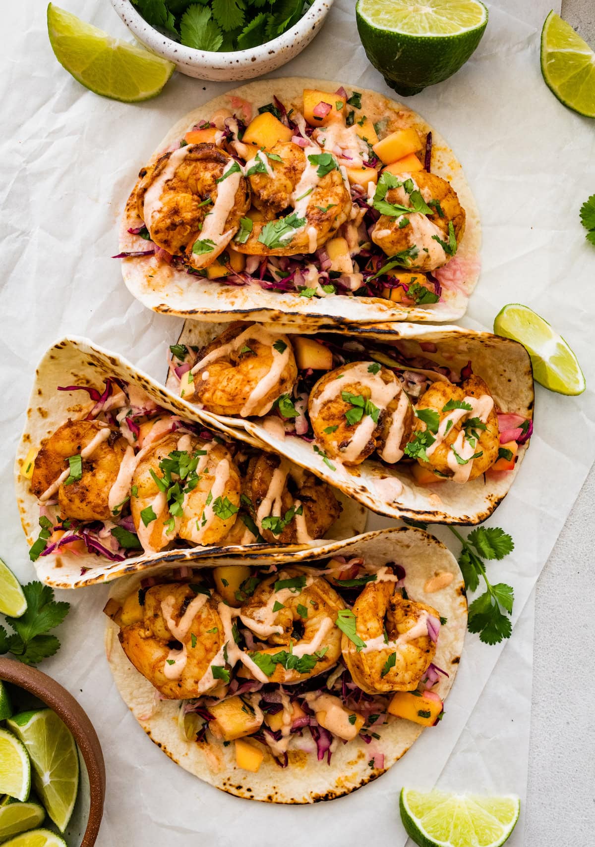 Four shrimp tacos in flour tortillas folded up. The tacos are topped with a mango cabbage slaw and a drizzle of spicy crema.