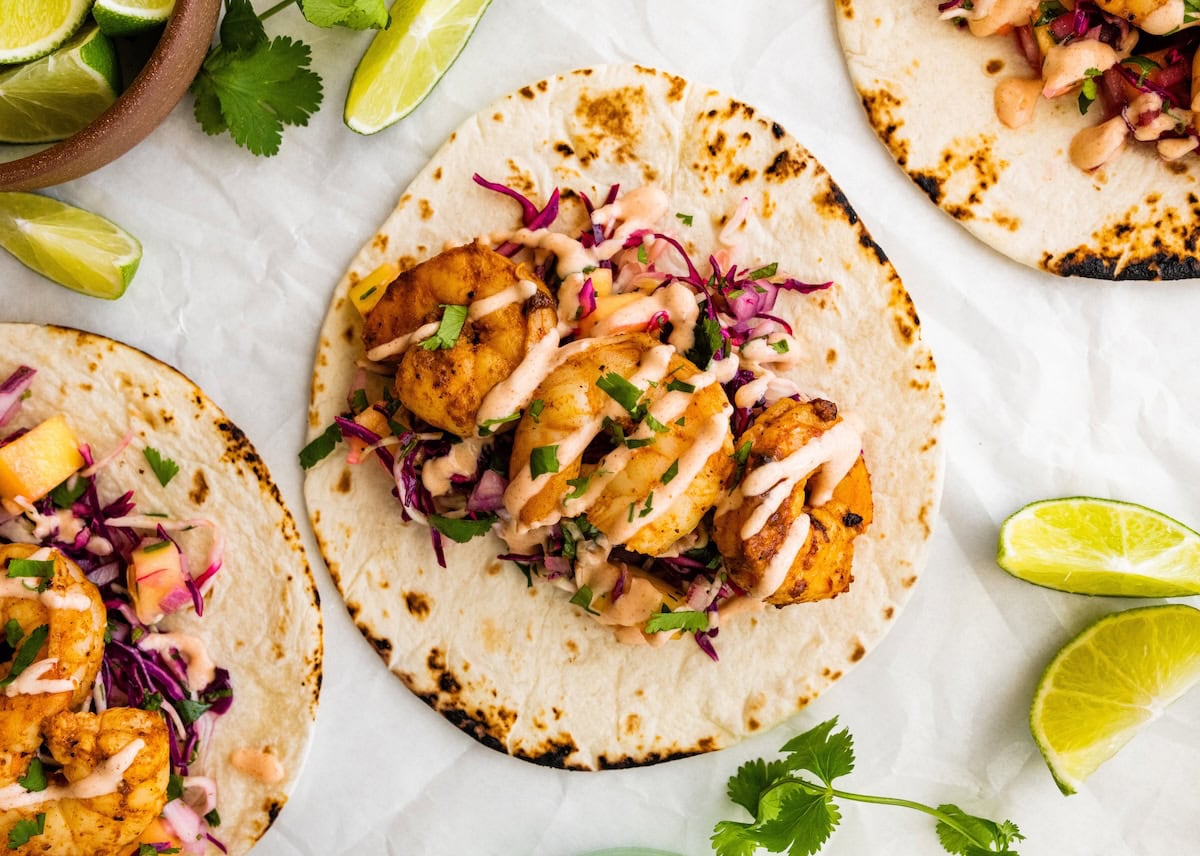 An open-faced shrimp taco on a flour tortilla. The shrimp taco is topped with mango cabbage slaw and a drizzle of spicy crema.