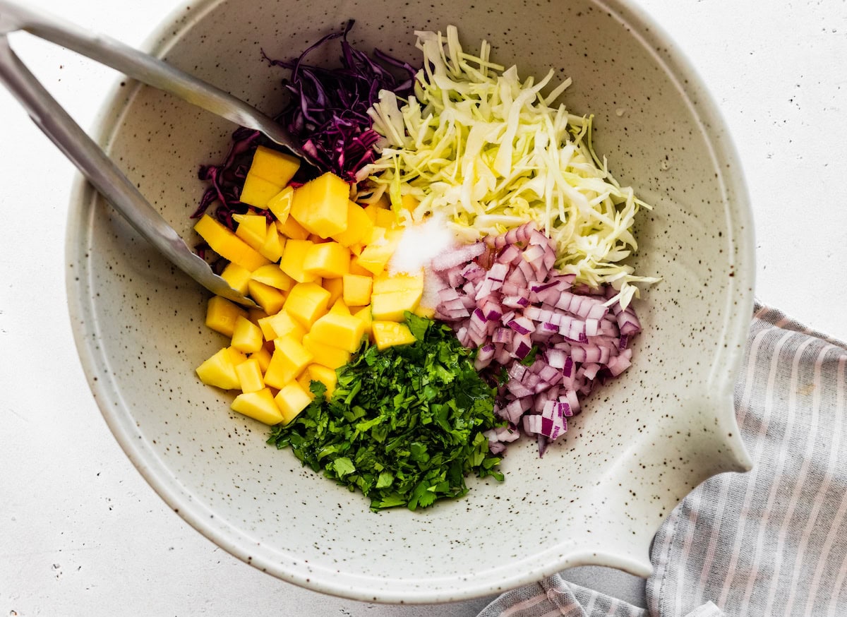The ingredients for the mango cabbage slaw in a mixing bowl with metal tongs: mango, cilantro, red onion, red cabbage and green cabbage.