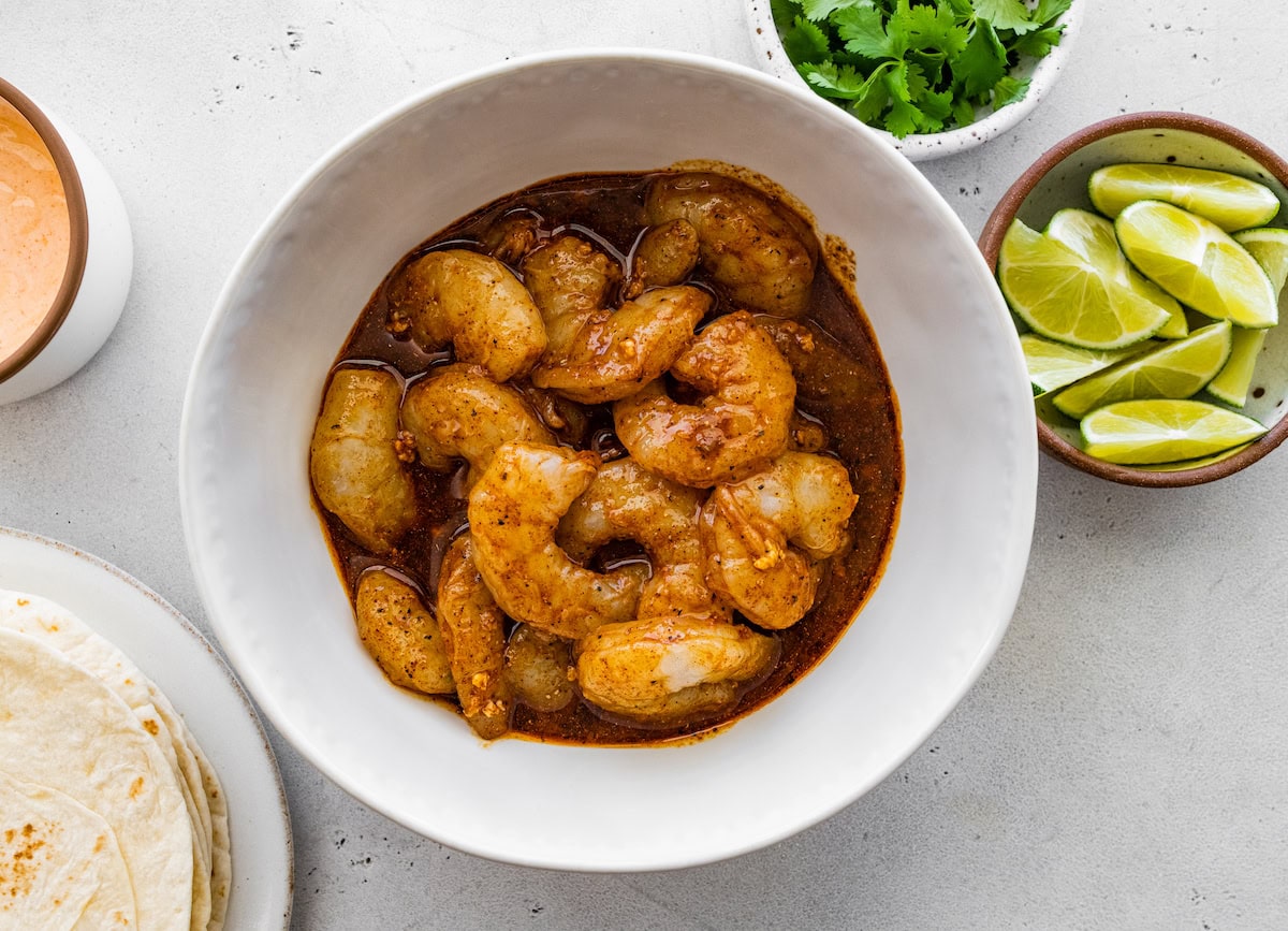 Raw shrimp marinating in a sauce in a small bowl. There is a small bowl with limes next to the shrimp.