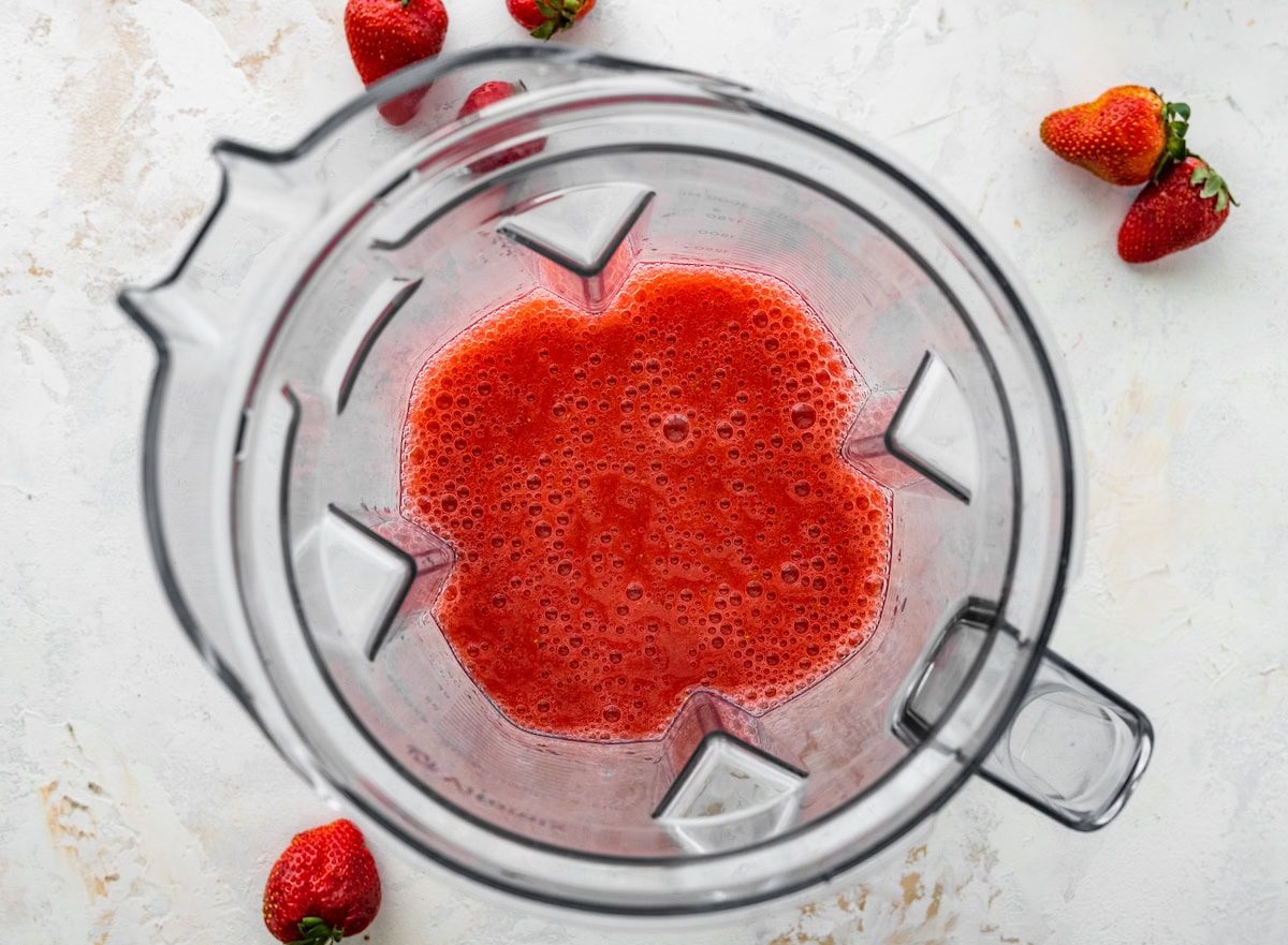 Freshly blended strawberry puree inside a blender, surrounded by scattered strawberries.
