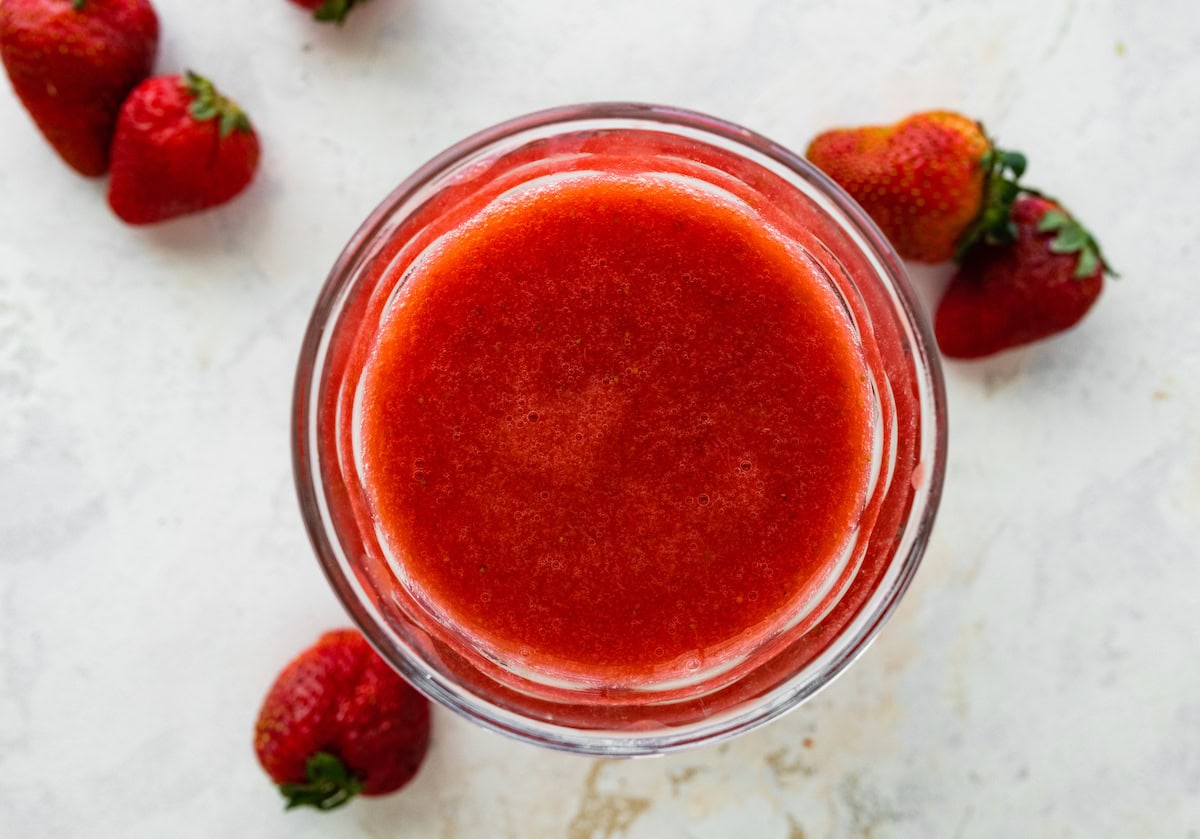 A glass jar filled with smooth strawberry juice, surrounded by whole strawberries.