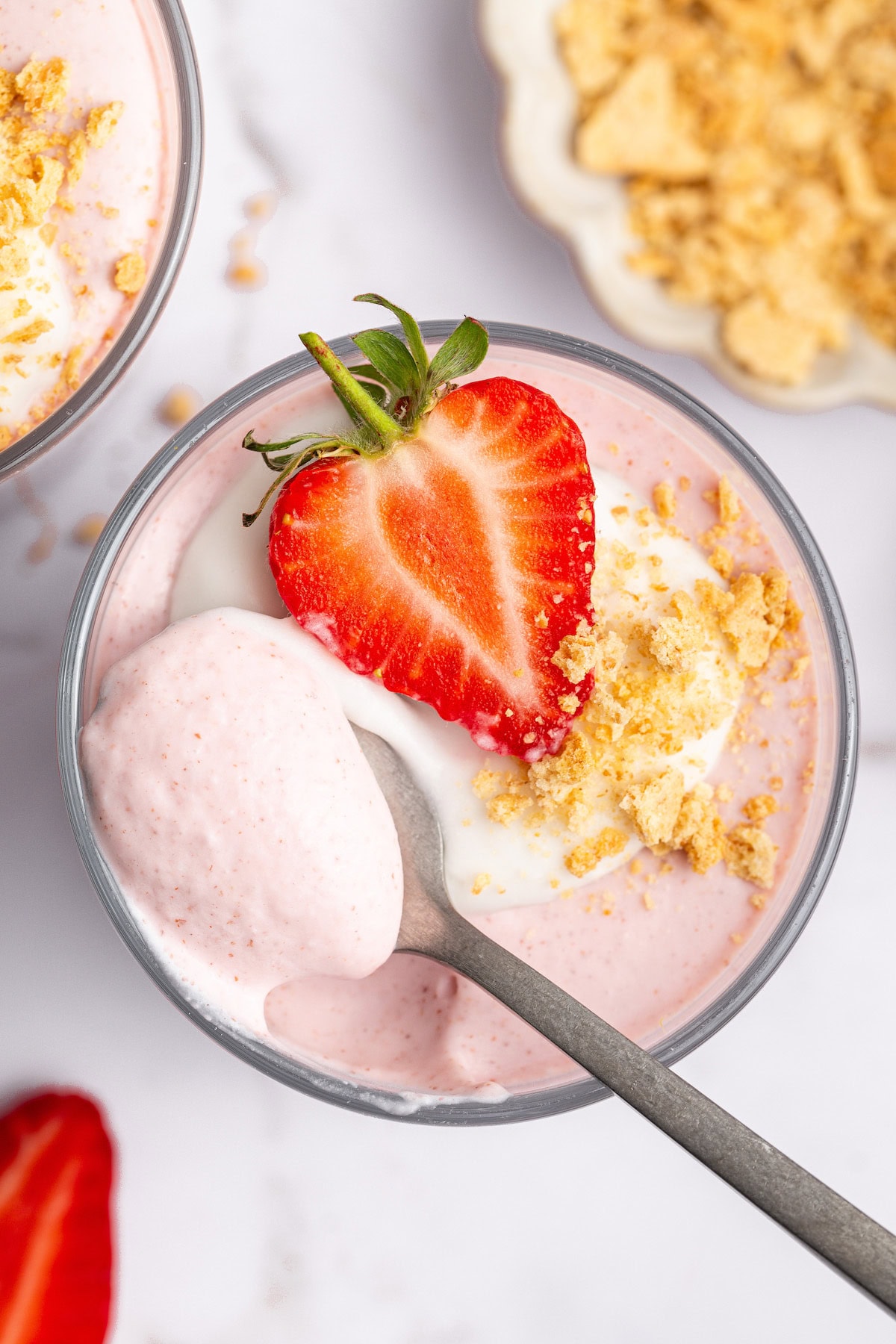 One glass filled with high-protein strawberry mousse topped with a dollop of cocowhip, one halved fresh strawberry and crushed graham crackers. There is a spoon scooping out a bite from the glass.