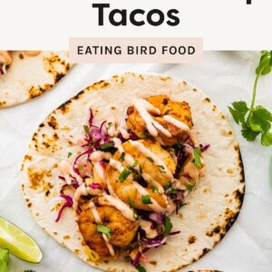 Grilled shrimp tacos topped with shredded cabbage, chopped cilantro, and a creamy drizzle, served on a toasted flour tortilla with lime wedges and fresh herbs on the side.