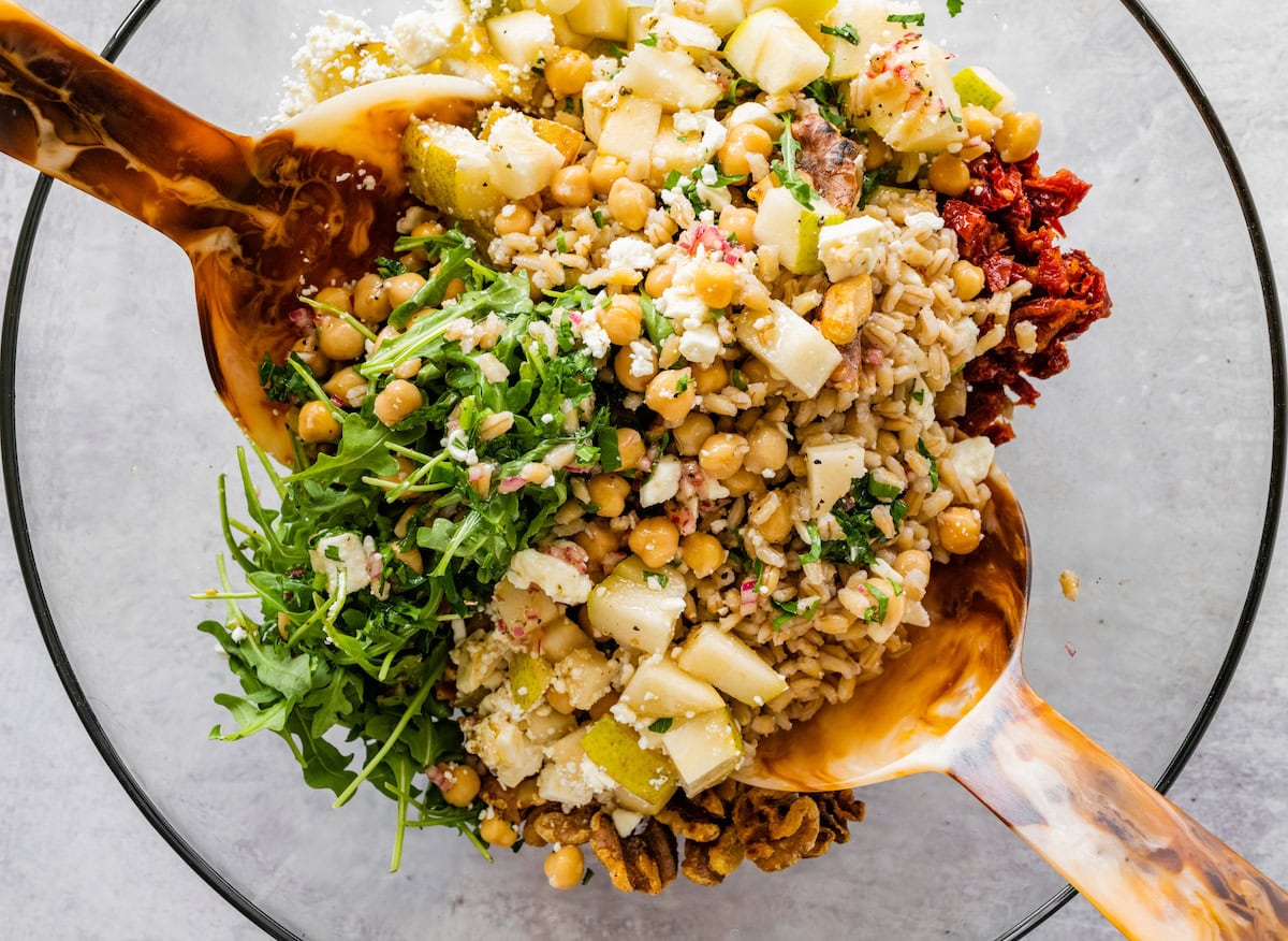 Barley salad in a large glass mixing bowl with two serving wooden spoons mixing all the ingredients.
