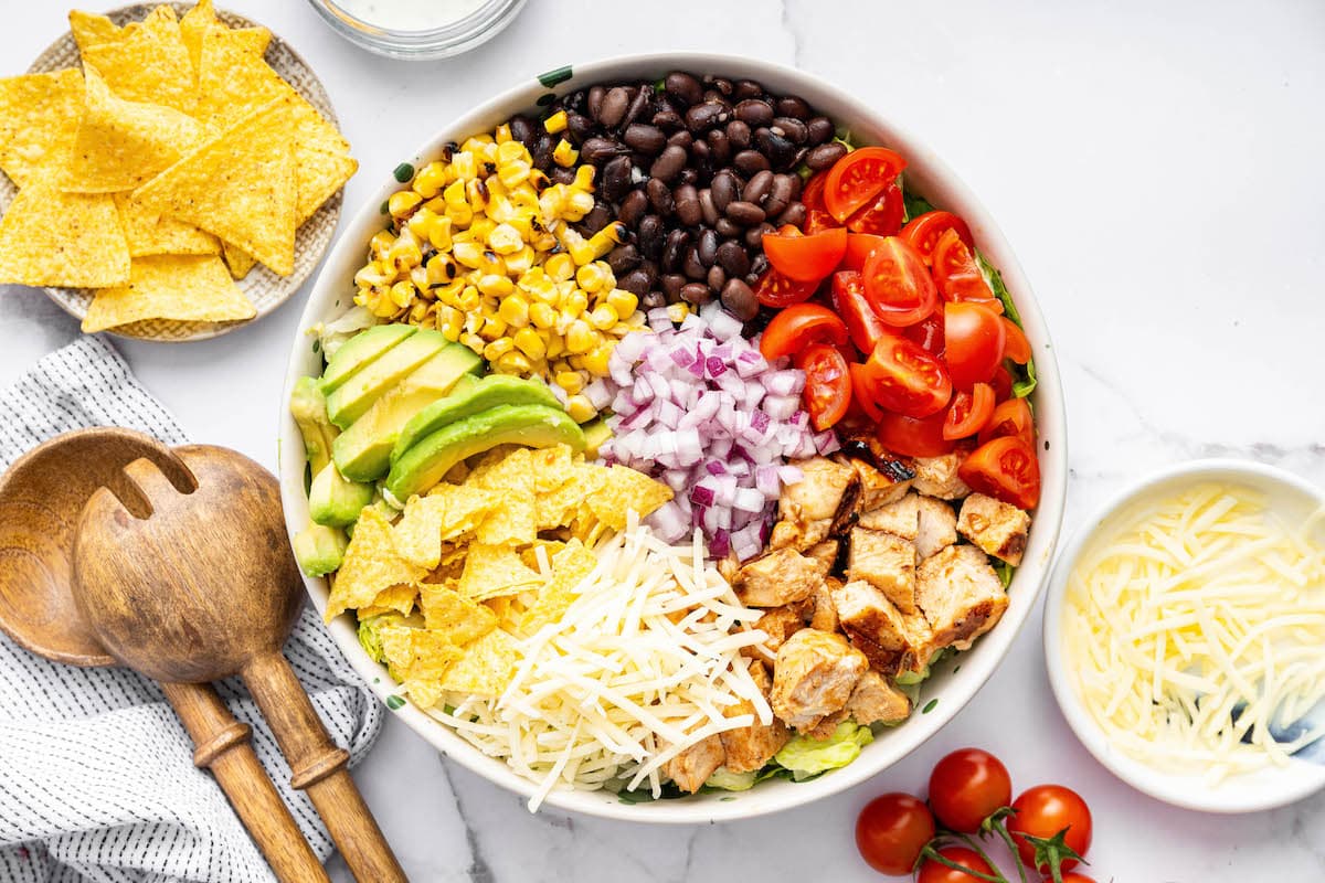 Ingredients for the BBQ chicken salad sectioned off on top of lettuce: red onions, black beans, cherry tomatoes, BBQ chicken, shredded cheese, avocado, corn and tortilla chips.