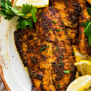 Blackened tilapia filets on a plate topped with fresh parsley and served with lemon wedges.