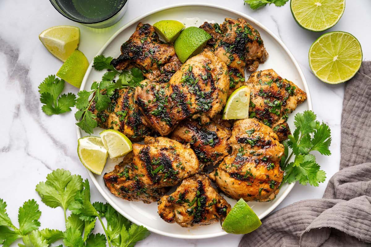 Multiple grilled chicken thighs on a plate, garnished with fresh cilantro and lime wedges to the side.