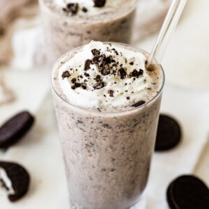 Two cookies and cream protein shakes in glasses, topped with whipped cream and crushed chocolate sandwich cookies, with clear straws inserted.
