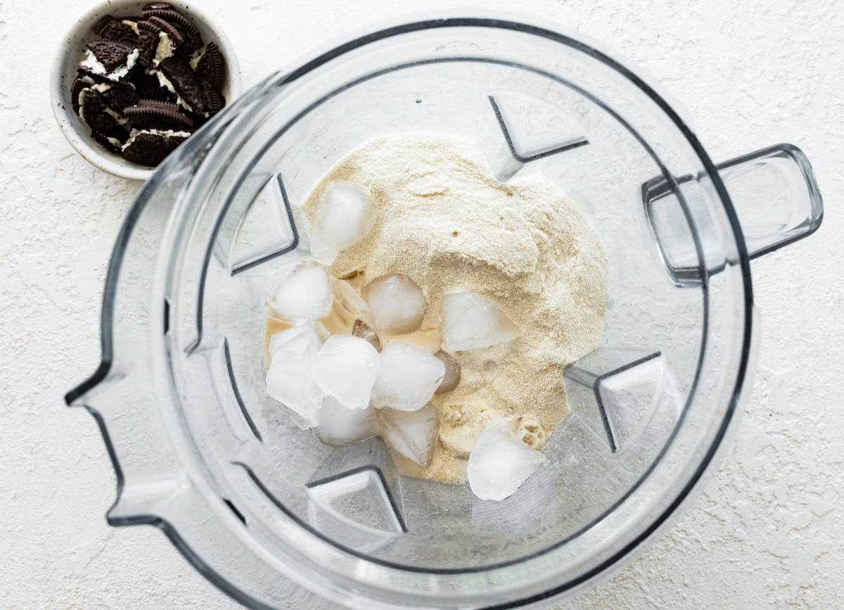 A blender containing ice cubes and vanilla protein powder, with a bowl of halved chocolate sandwich cookies next to it.