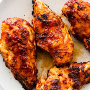 Four grilled BBQ chicken breasts on a white serving plate.